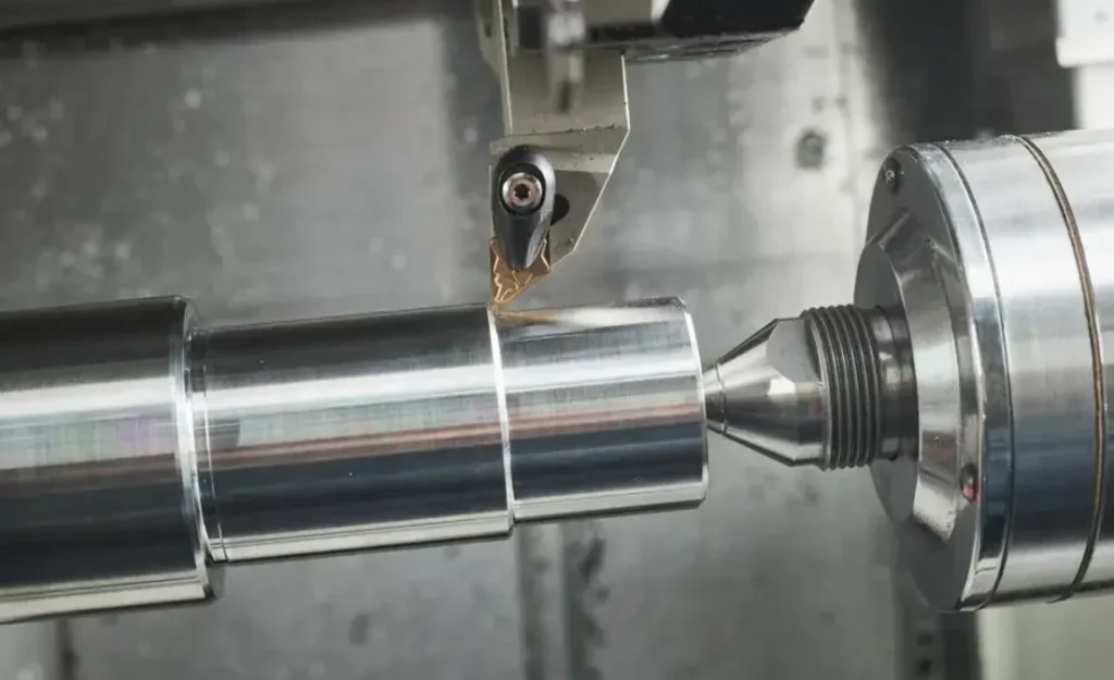 Metal shaft being machined on a CNC lathe; the cylindrical workpiece is rotating while a visible cutting tool is in contact with its surface