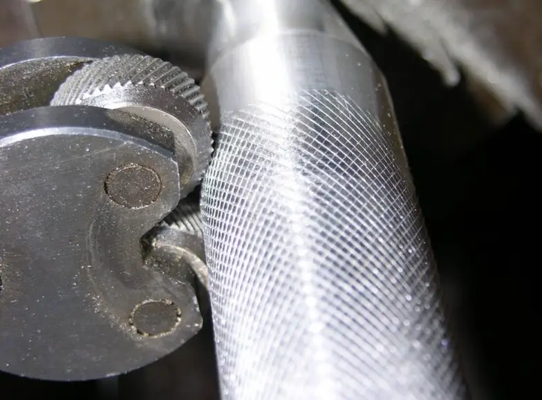 Close-up of a cylindrical metal part with serrated knurling patterns being formed by a knurling tool featuring two opposing wheels