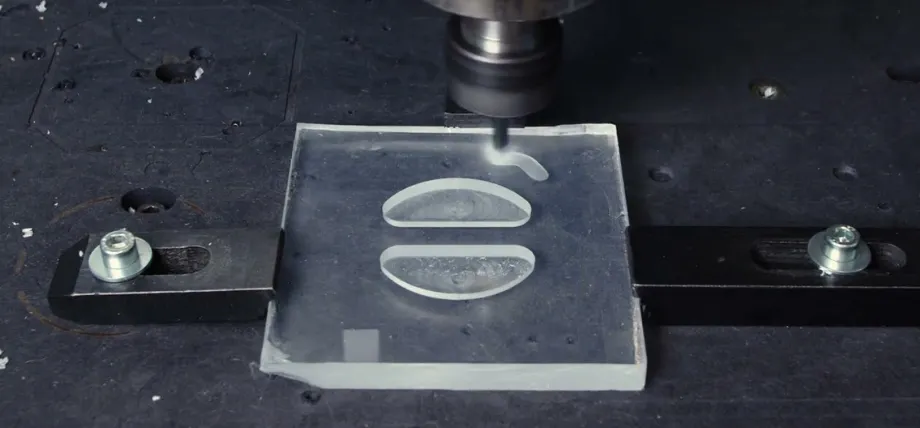 A CNC spindle is performing a milling operation on a clear acrylic sheet, using a high-speed bit to carve out precise geometric shapes.