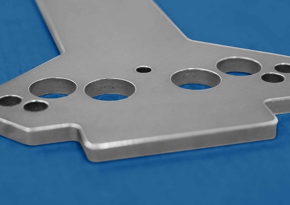 Laser-cut stainless steel flat part with smooth deburred edges and multiple circular holes on a blue surface.