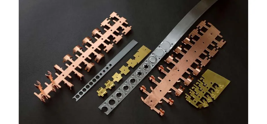 Various metal strips in copper, brass, and steel showing different stages of the progressive die stamping process.