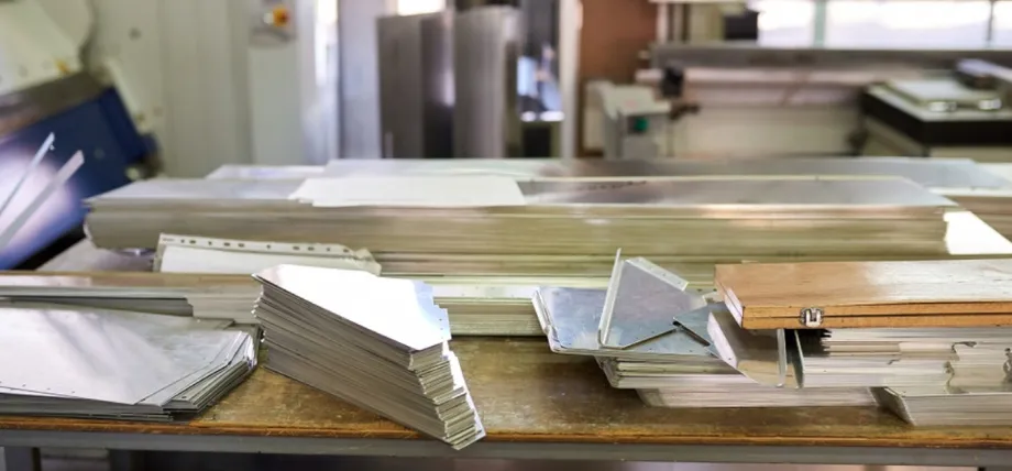 Organized stacks of cut and formed aluminum sheet metal pieces, including flat sheets, angled brackets, and perforated components
