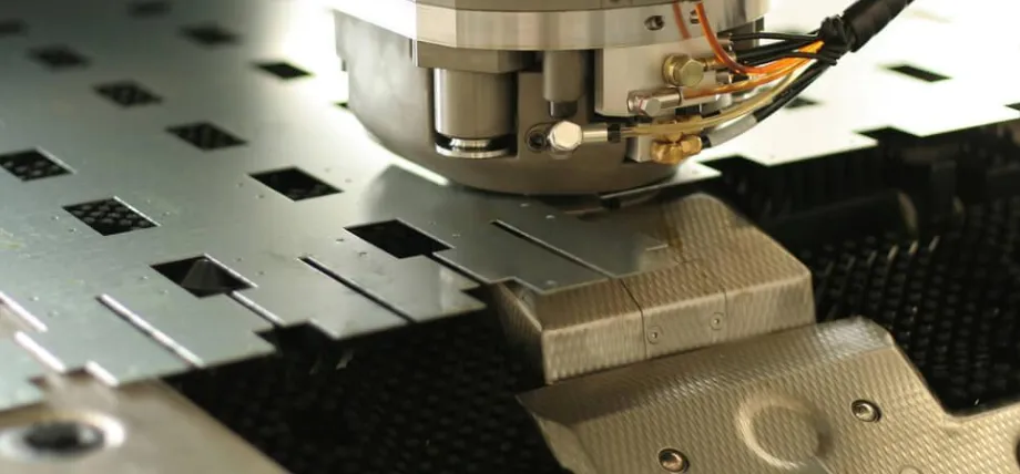 A close-up of a CNC punching machine head perforating a grey metal sheet with precise rectangular cutouts.