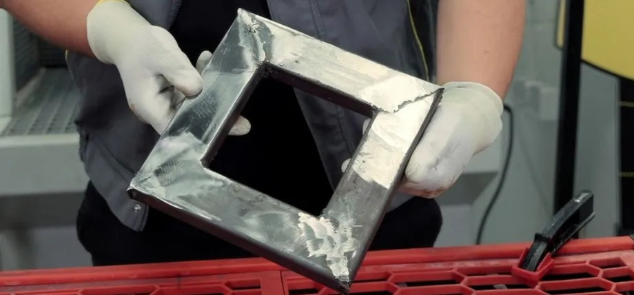 worker wearing gloves and protective clothing holds up a freshly welded square metal frame fabricated from sheet steel