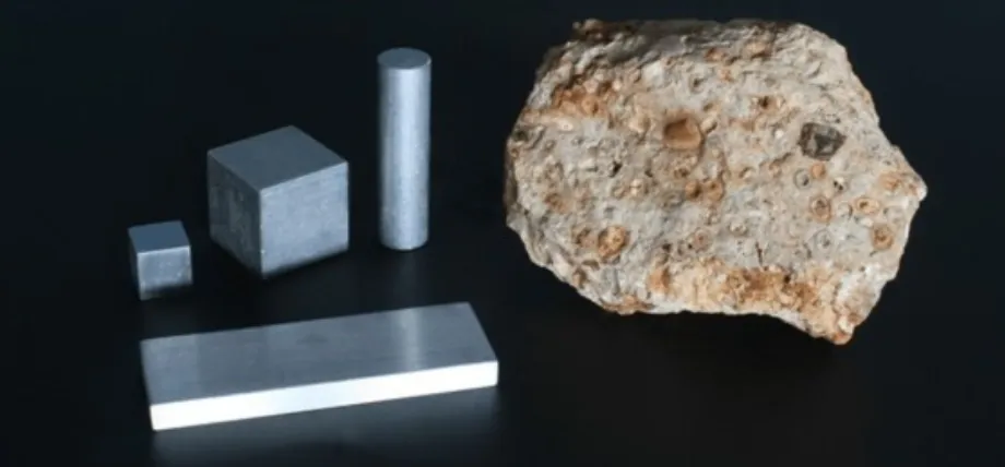 A large piece of raw bauxite ore shown alongside processed aluminum samples in cube, cylinder, and plate forms.