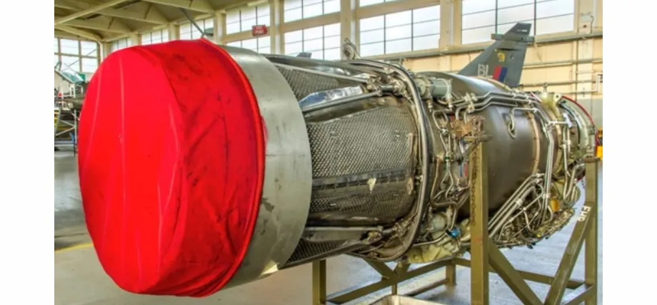 A close-up of a complex aerospace turbine engine mounted on a stand, featuring intricate piping and a large red protective intake cover.
