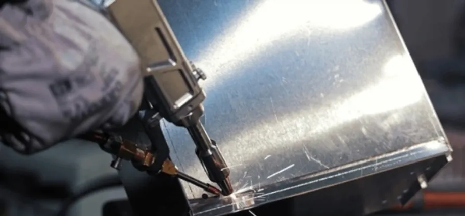 A worker using a handheld spot welding tool on a shiny metal box, with small sparks emitting from the localized heat point
