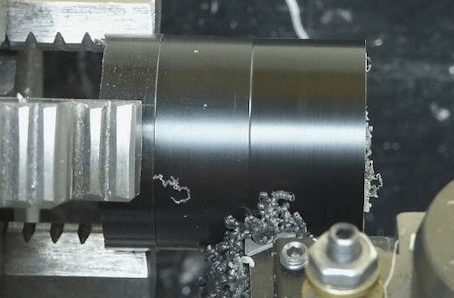 CNC lathe turning a black acetal rod with curled plastic chips accumulating at the cutting tool. 