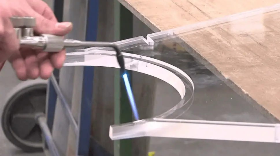 A small torch is being used to flame-polish the edge of a curved, clear polycarbonate part.