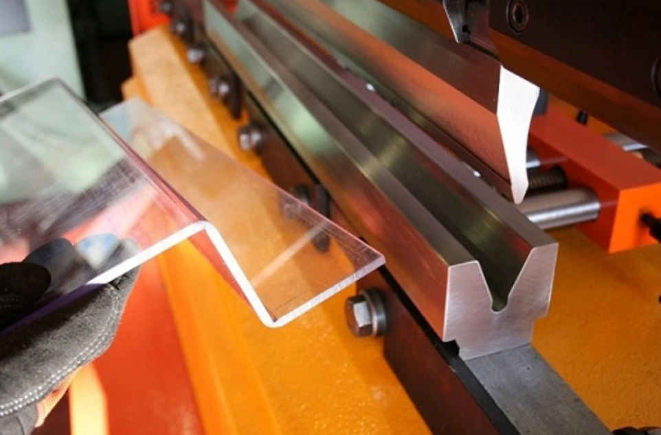 Gloved hand holding a Z-shaped bent polycarbonate sheet on an orange press brake machine.