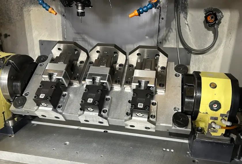 Multiple rigid precision vise fixtures mounted on a CNC machine table with coolant nozzles and a rotary axis