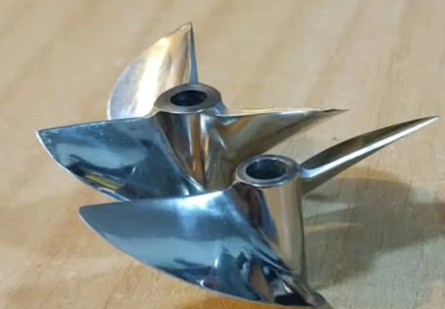Two polished CNC-machined titanium boat propellers with twisted blades and center bores, placed on a surface. 