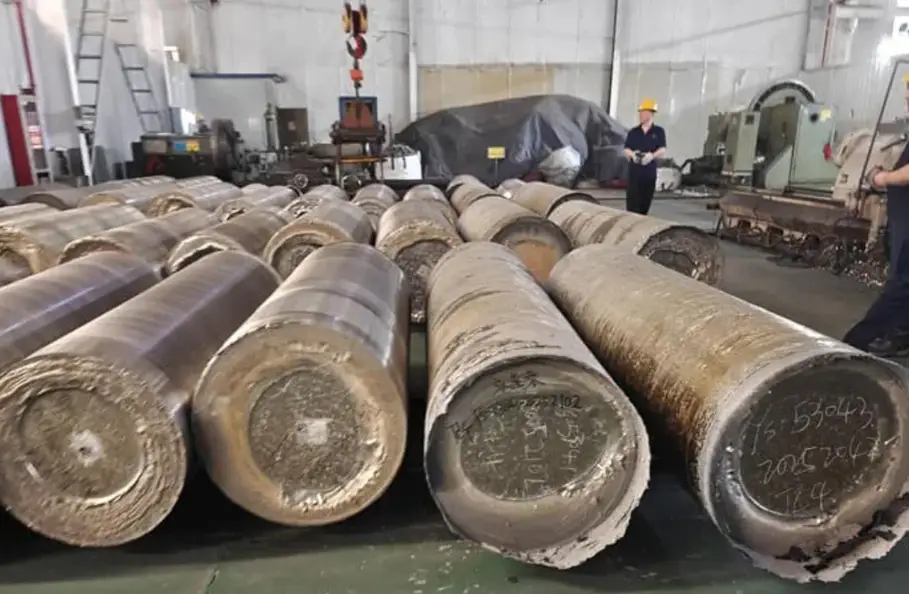 Large raw titanium billets with mill markings stored on a factory floor, with industrial machinery and workers in the background