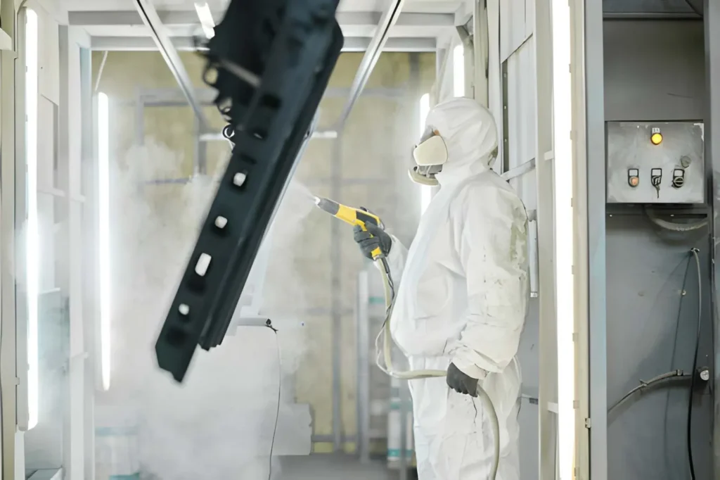 Worker applying powder coating to metal using a spray gun in a factory.