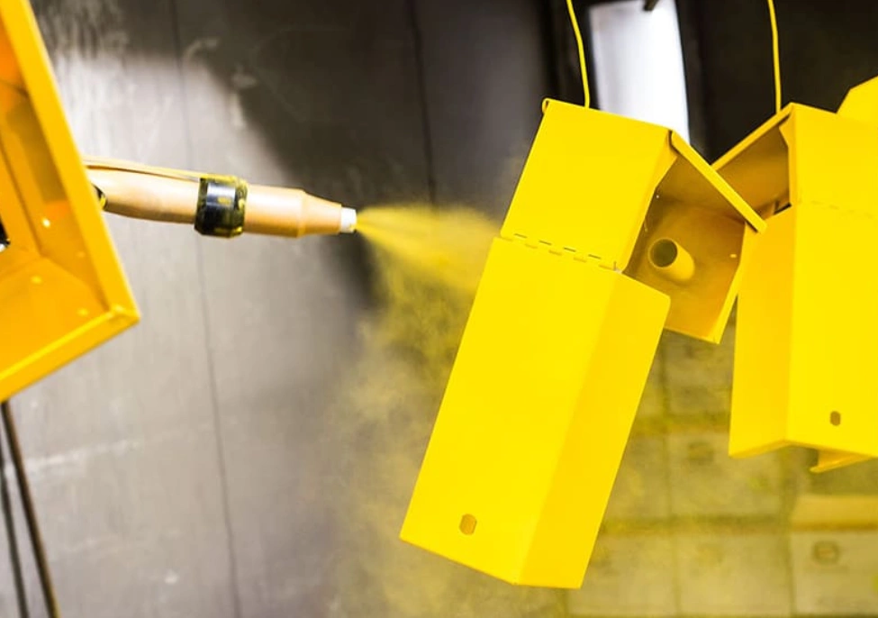 Powder coating gun applying yellow finish to suspended laser-cut brass metal parts for surface protection.
