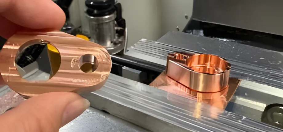 A machinist holding a freshly milled copper component over a CNC machine bed, displaying a high-shine reddish-orange surface.