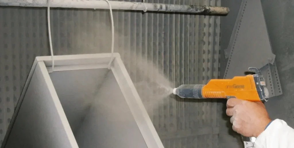 Person applying powder coating to a metal component using an electrostatic spray gun in an industrial coating booth.