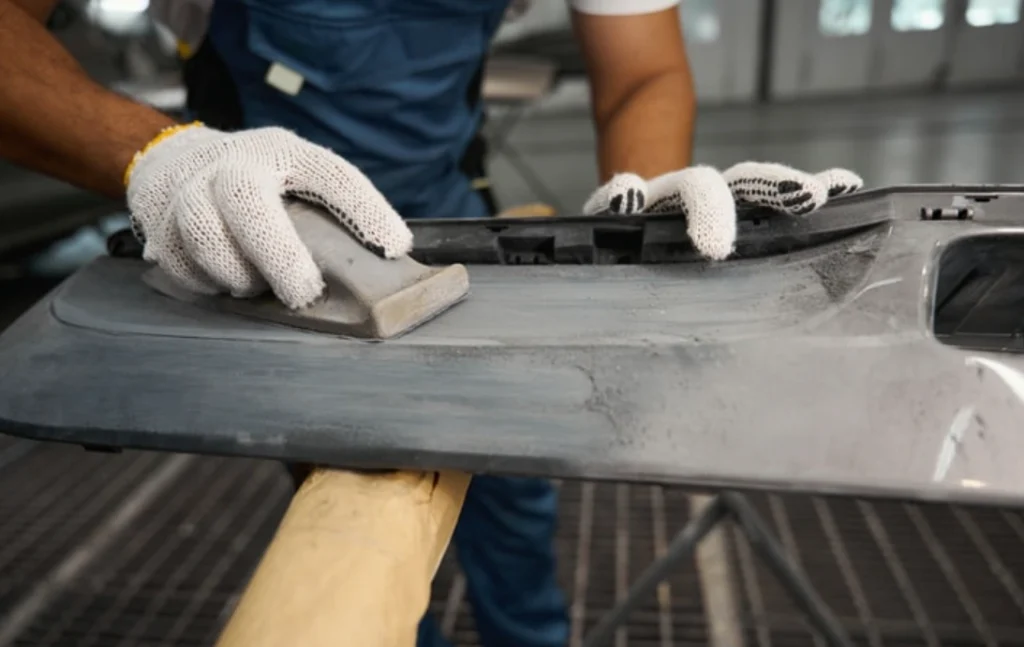 Gloved worker hand-sanding a metal automotive panel during surface preparation before coating