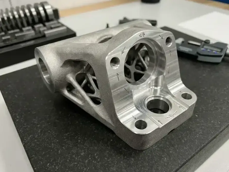 Custom aerospace bracket with lattice supports and machined mounting faces beside a digital caliper.